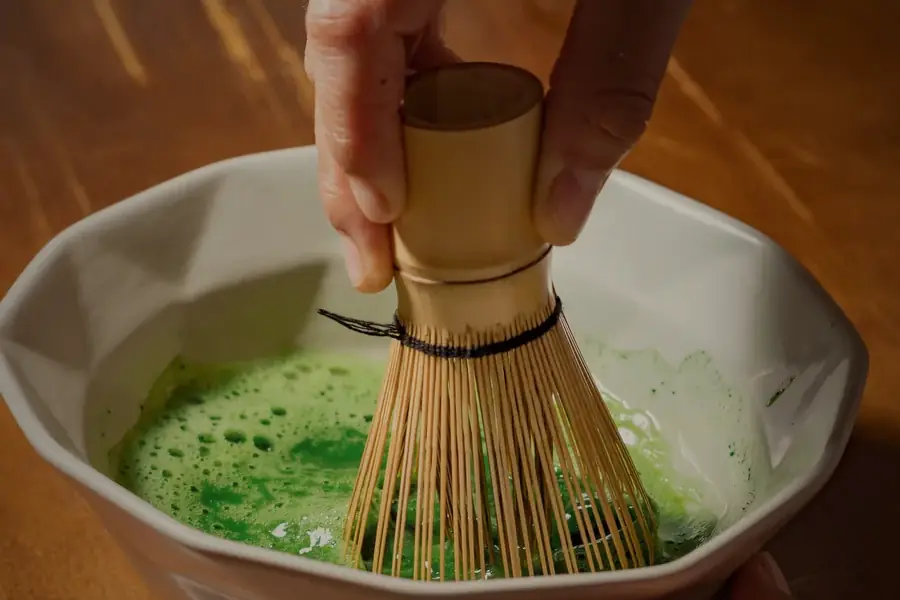 Beyaz kâsede çasenle çırpılan matcha, sıcak ahşap masa.