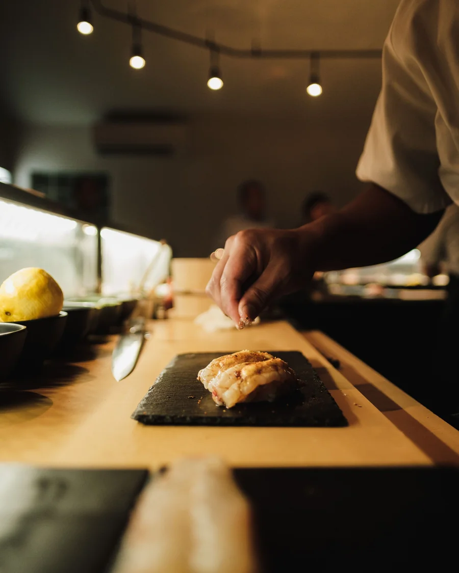 Hinoki tezgâhta arduvaz üstünde tek nigiri; ustanın eli üstüne tuz serpiyor, yanda narenciye.